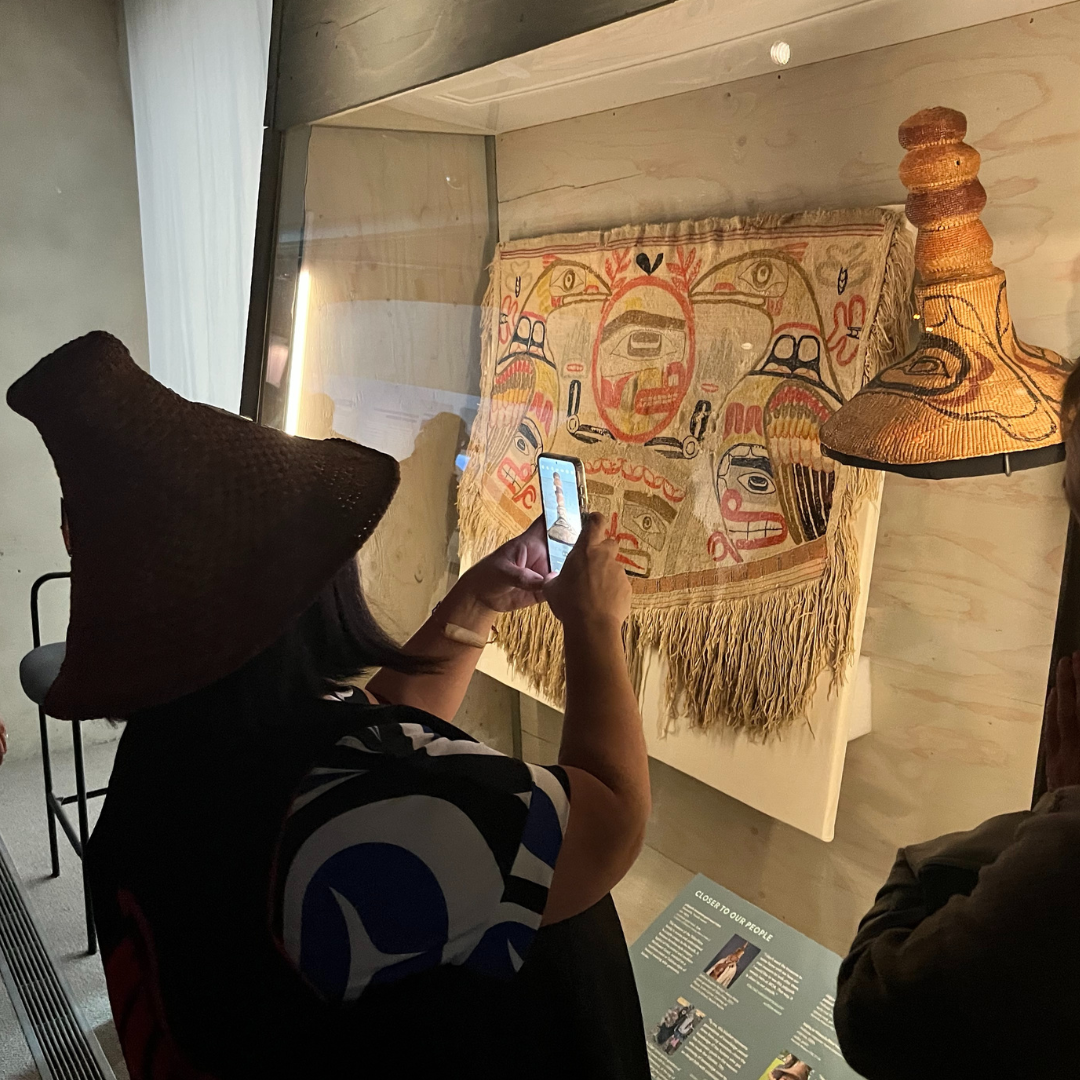 An individual holds up their phone to take a photo of a potlatch hat in a museum display case. The individual is wearing a similarly shaped hat.