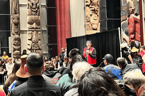 An individual stands at the front of a large room in front of a black backdrop, speaking into a microphone to a large crowd of people. The exterior of the room displays large artworks.