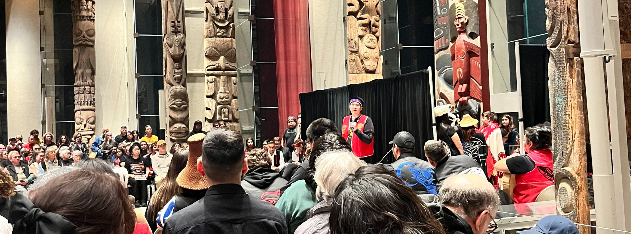 An individual stands at the front of a large room in front of a black backdrop, speaking into a microphone to a large crowd of people. The exterior of the room displays large artworks.