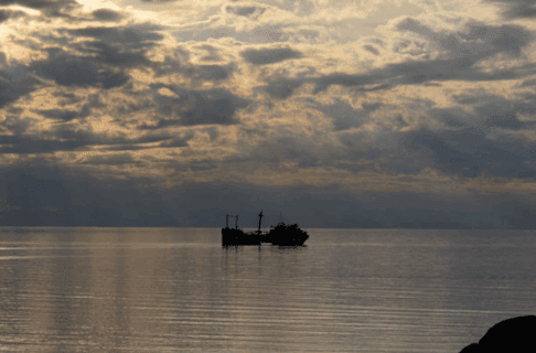Looking out over a body of water towards a partially emerged shipwreck lit by sunlight breaking through dark clouds.