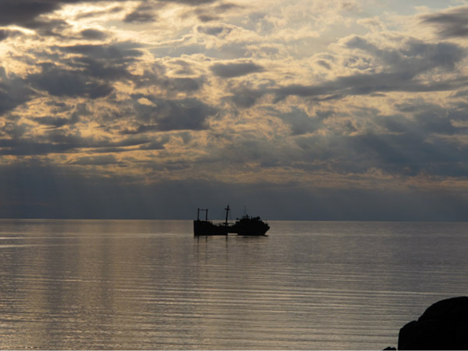 Looking out over a body of water towards a partially emerged shipwreck lit by sunlight breaking through dark clouds.