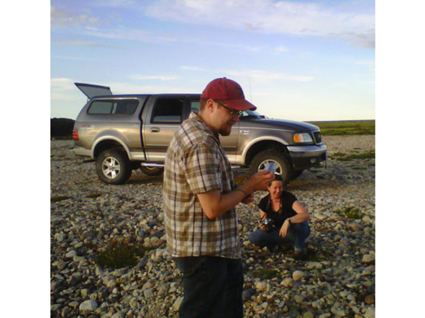 An individual crouches down near the rocky ground holding a camera. Behind them a truck with a covered bed is parked with the back hatch open, and in the foreground is a standing individual wearing a red baseball cap.