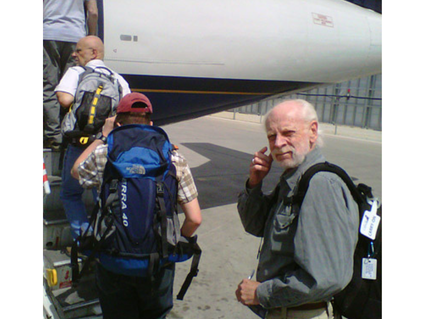 Three people wearing backpacks boarding a plane from the tarmac. The last person in line turns to look back towards the photographer.