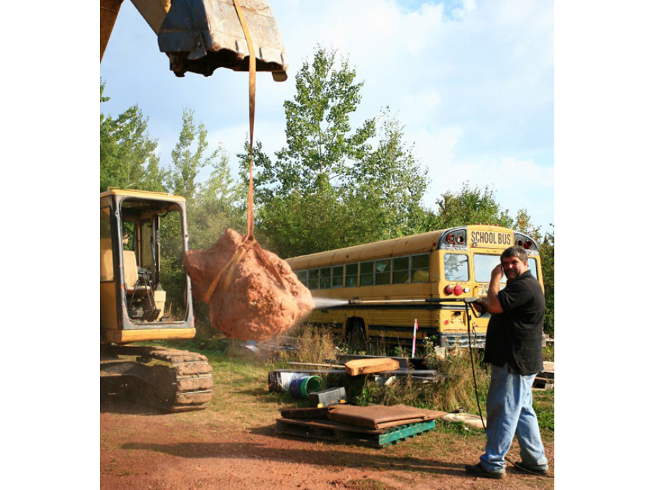 A large boulder suspended by the scoop of a bulldozer as a person pressure washes it.