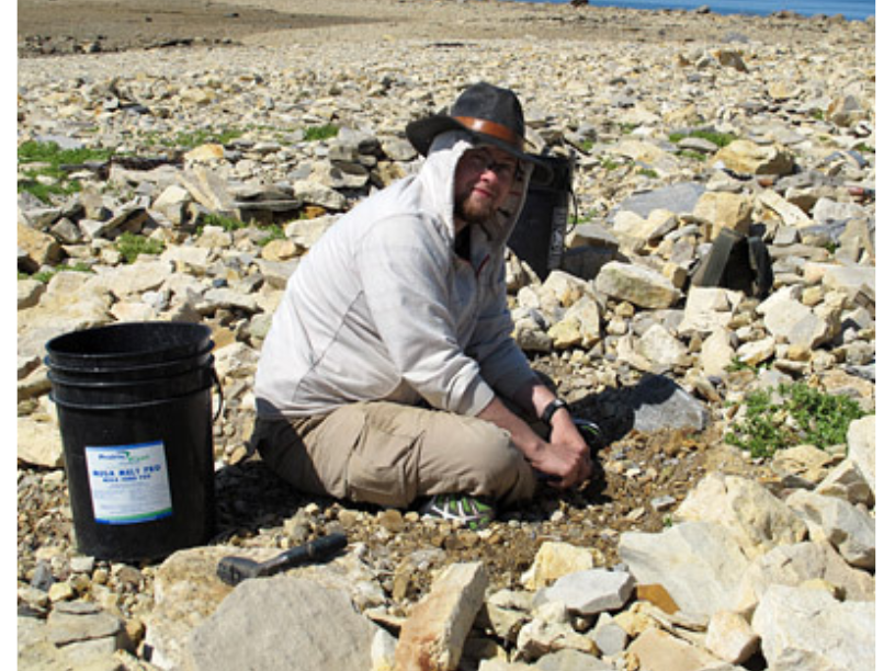 An individual seated crossed-legged on a rocky surface next to a black bucket. They have the hood of their top up, and a rimmed hat.