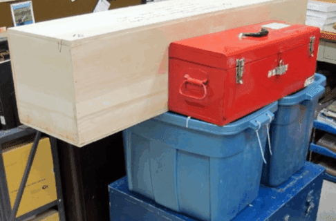 A stack of boxed supplies packed for transport. At the base a large blue crate, with two blue rubber bins on top of it. Topping off the pile, a long light-coloured wood crate and a red toolbox.