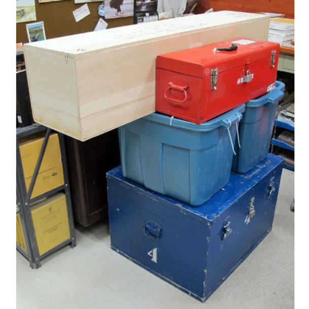 A stack of boxed supplies packed for transport. At the base a large blue crate, with two blue rubber bins on top of it. Topping off the pile, a long light-coloured wood crate and a red toolbox.