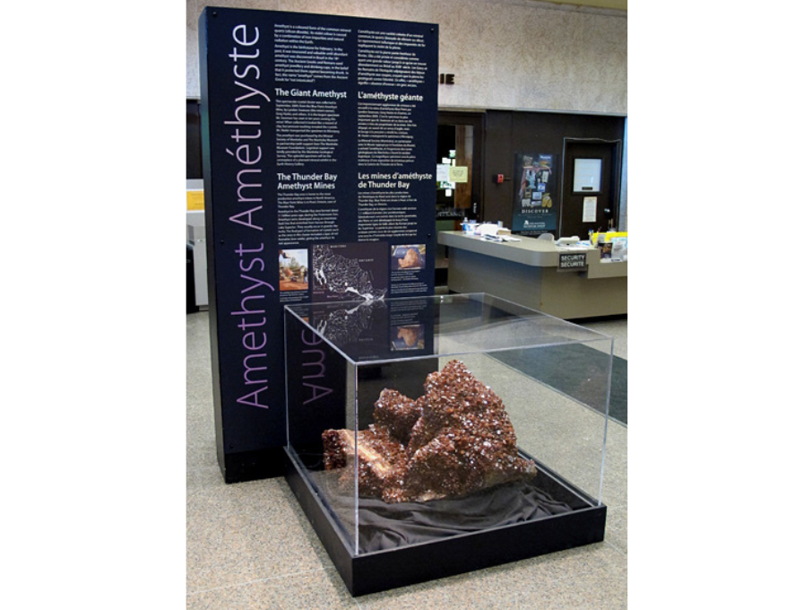 A large amethyst in a plexiglass display case in front of a large information panel.