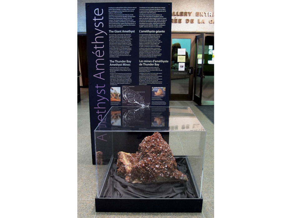 A large amethyst in a plexiglass display case in front of a large information panel.