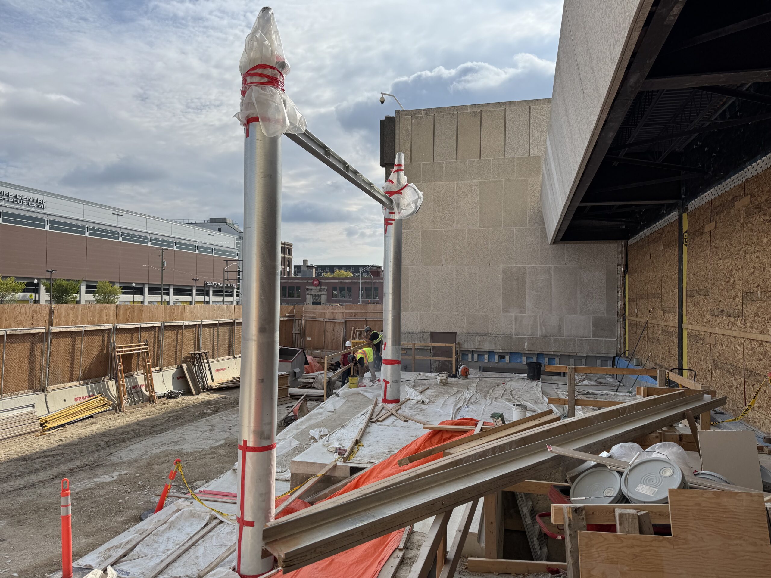 Rupert entrance to the Manitoba Museum under construction. Two tall pillars, connected by a beam near the top are errected in front of a zig-zag concrete pad for a accessible ramp.