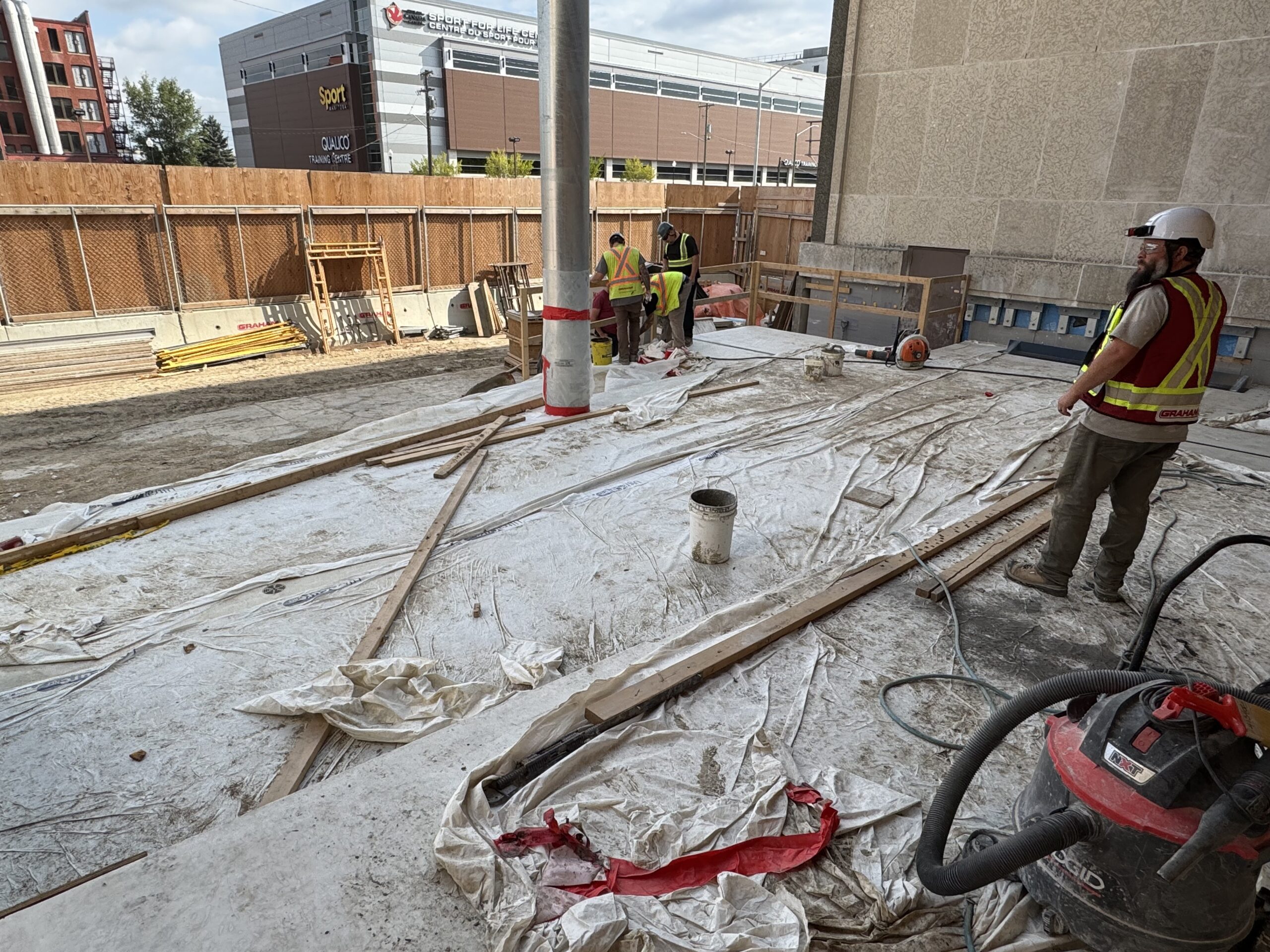 Looking out over a partially covered base for a concrete accessibility ramp leading up to the Manitoba Museum's Rupert Ave. doors. Several construction workers stand together near the lower portion working on a specific spot.