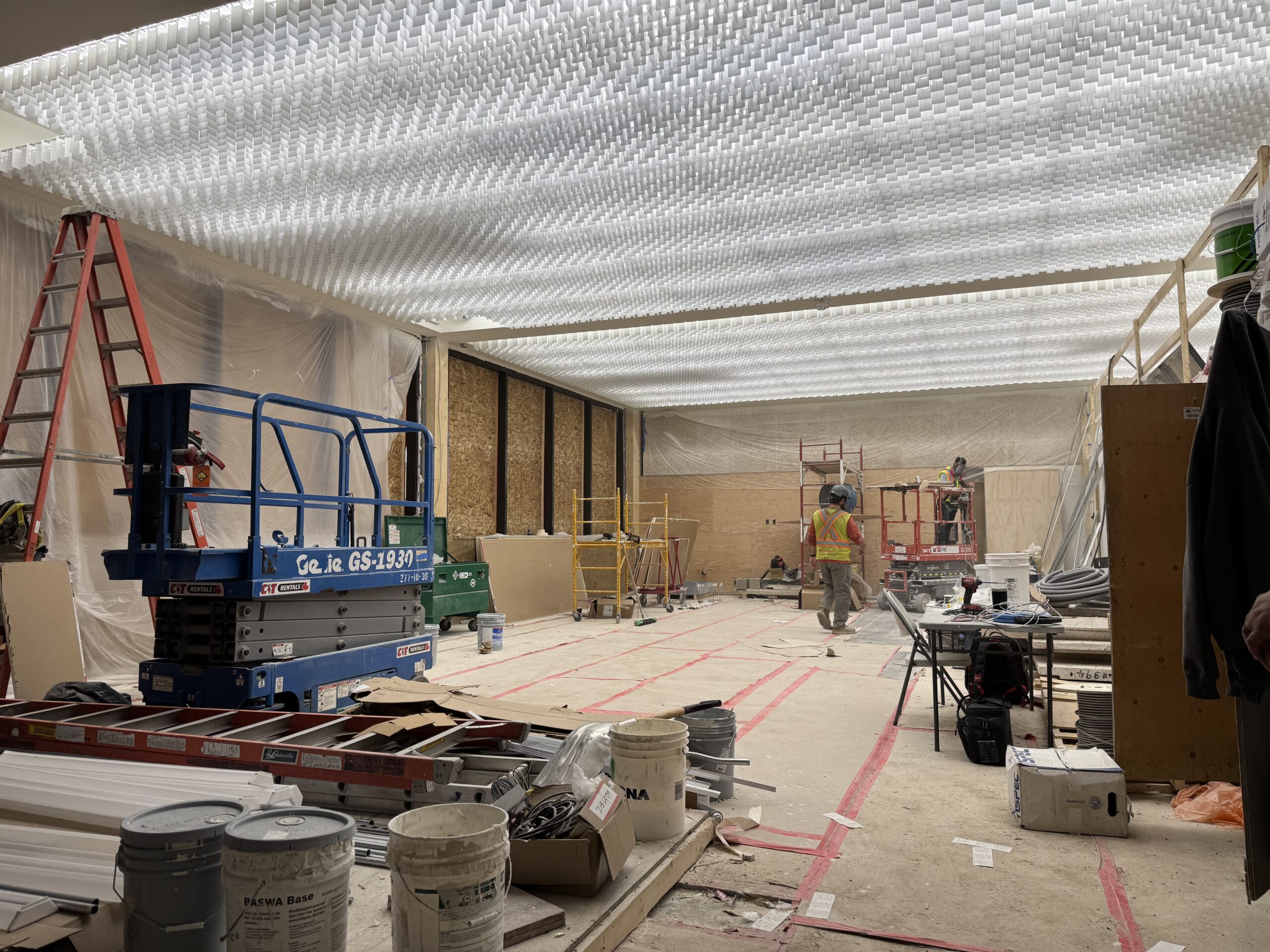 North side of the Museum foyer, under construction. Flooring is covered with protective materials, and two construction workers do work on the far side of the space with the aid of a scissor lift. New, bright white light covers adorn the ceiling.