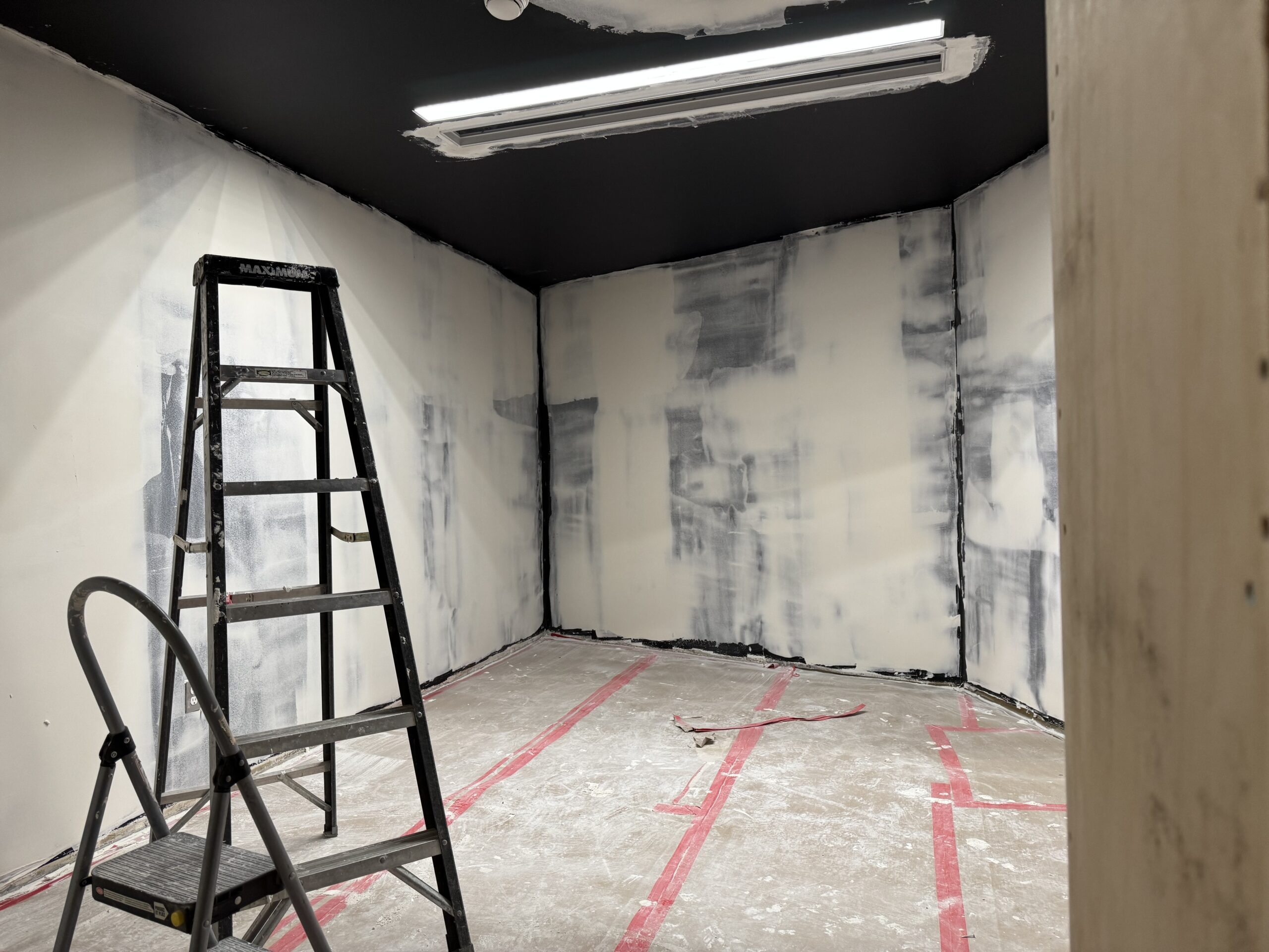 A small room constructed of drywall walls with a thin base coat of paint, and a clear tarp protected floor. A ladder stands in the lower left corner.