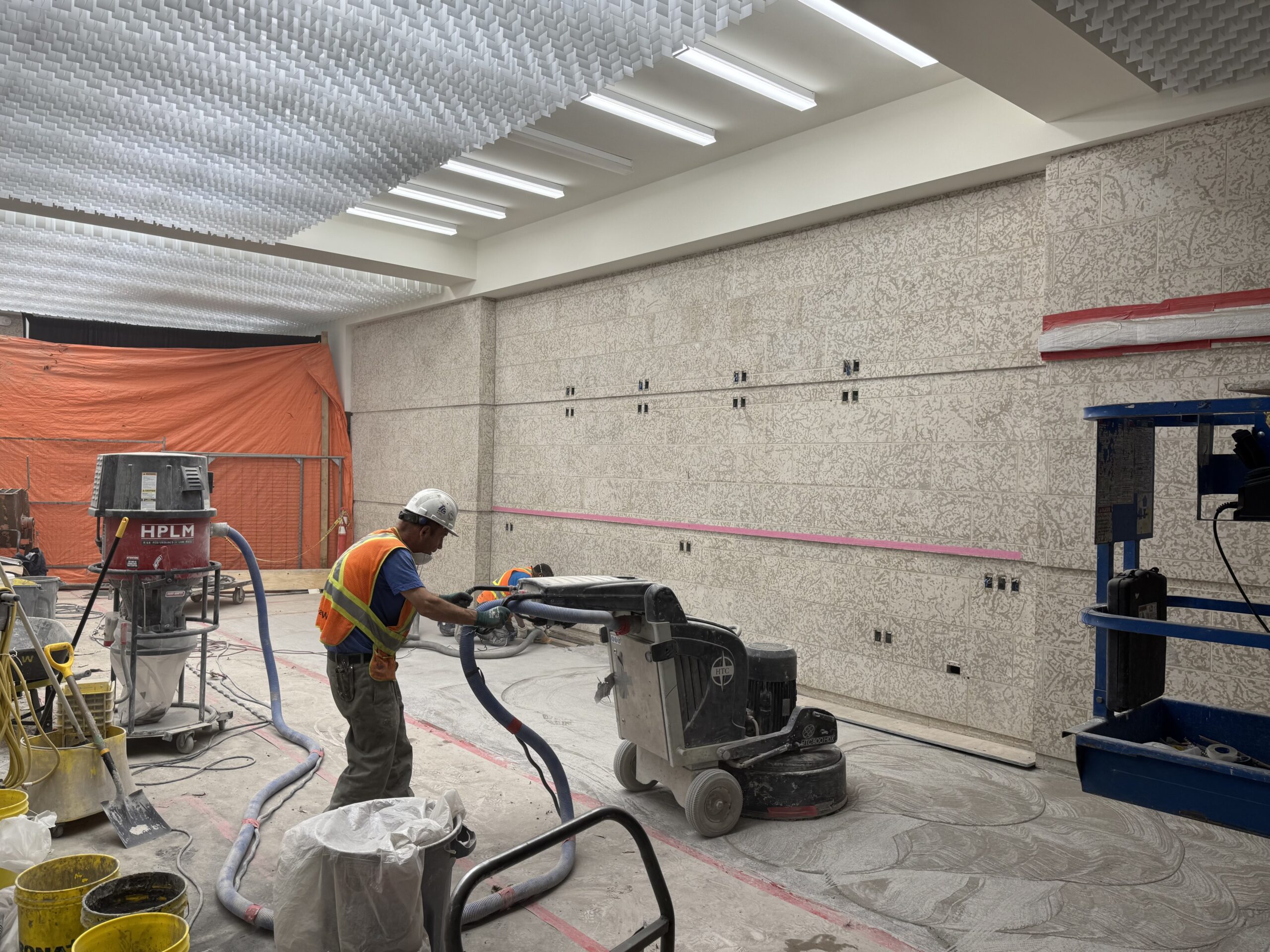 The south side of the Museum foyer under construction. A worker in PPE buffs a portion of the floor with a large machine, as another worker in the background works lower to the ground. Tech connections have been installed for the Box Office screens, and newly installed bright white light light covers adorn the ceiling.