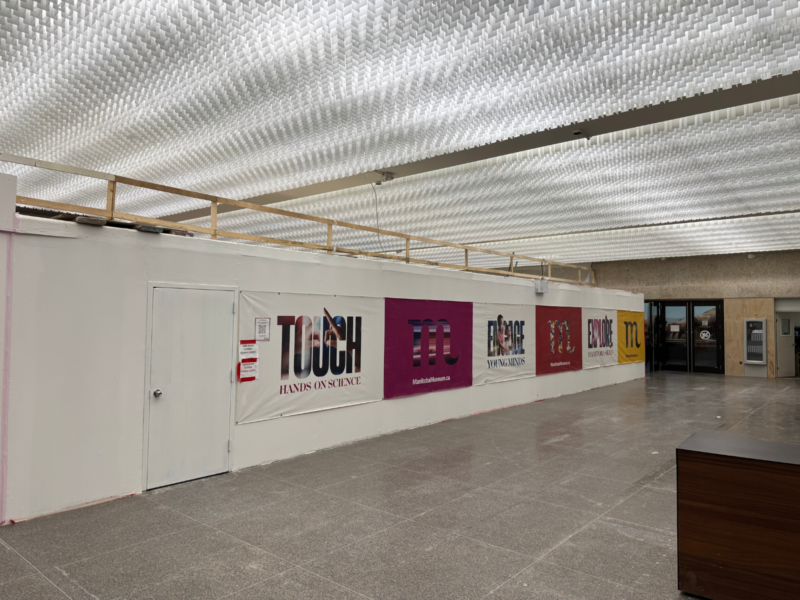 The Manitoba Musuem foyer, partially blocked off with temporary hoarding, but with a wide portion open leading to the Museum Galleries. On the hoarding wall is a banner with Museum branding.