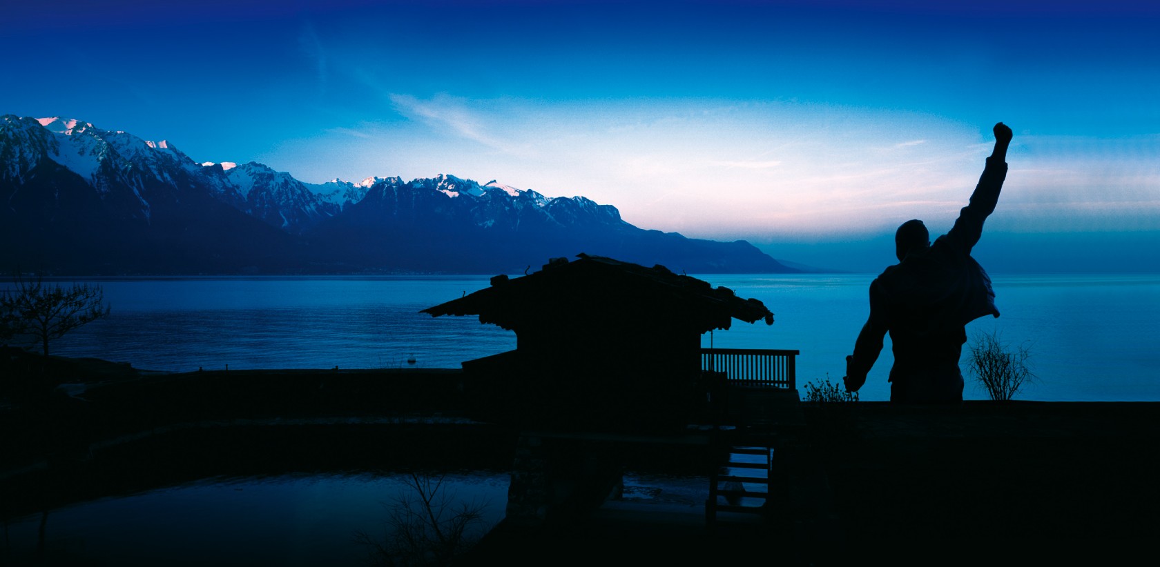 A silhouette of Freddie Mercury with his fist in the air standing on a dock in front of a lake surrounding by mountains.
