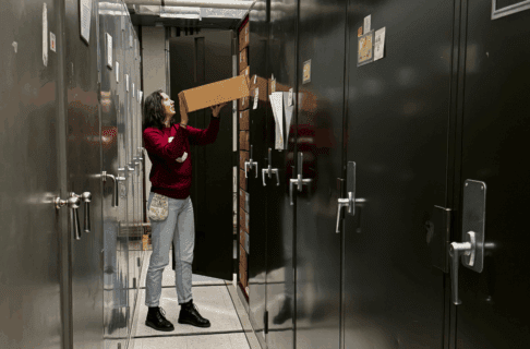 An individual in a red sweater and jeans standing between two rows of metal storage cabinets in front of an open cabinet. They are lifting a box of specimens back onto a shelf.