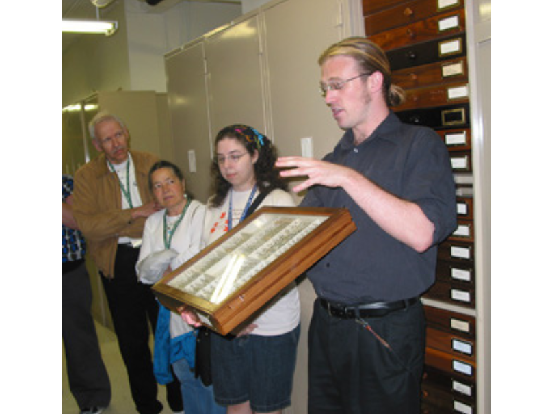 A museum staff person holding up a glass-covered museum storage drawer to a group of people.