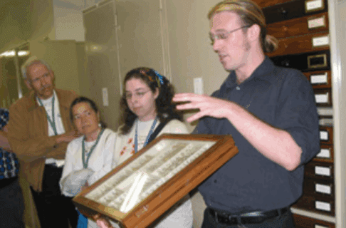 A museum staff person holding up a glass-covered museum storage drawer to a group of people.