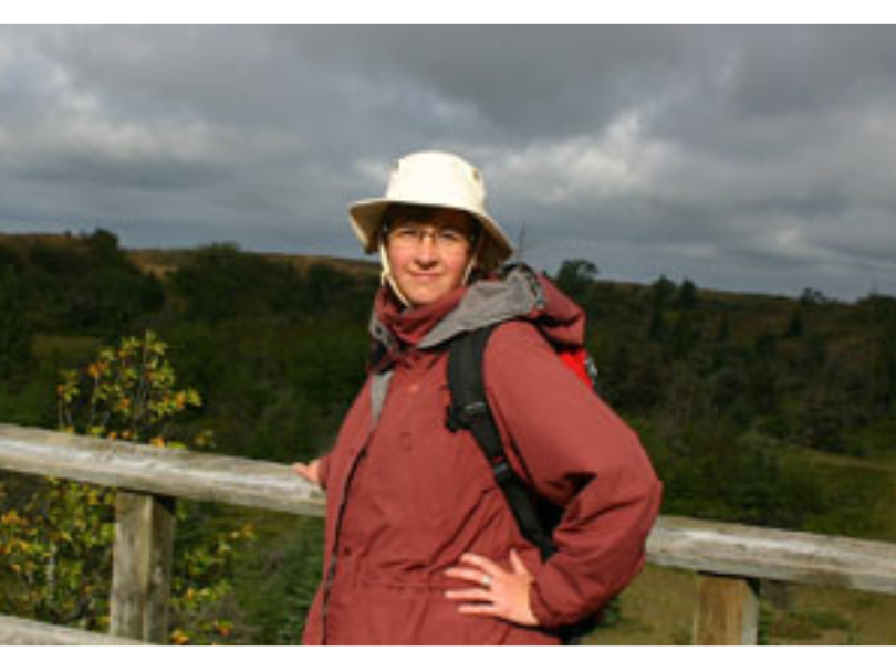 Dr. Diana Bizecki Robson wearing a rain jacket, wide-brimmed hat, and backpack standings at a wooden railing looking out over a treed landscape.