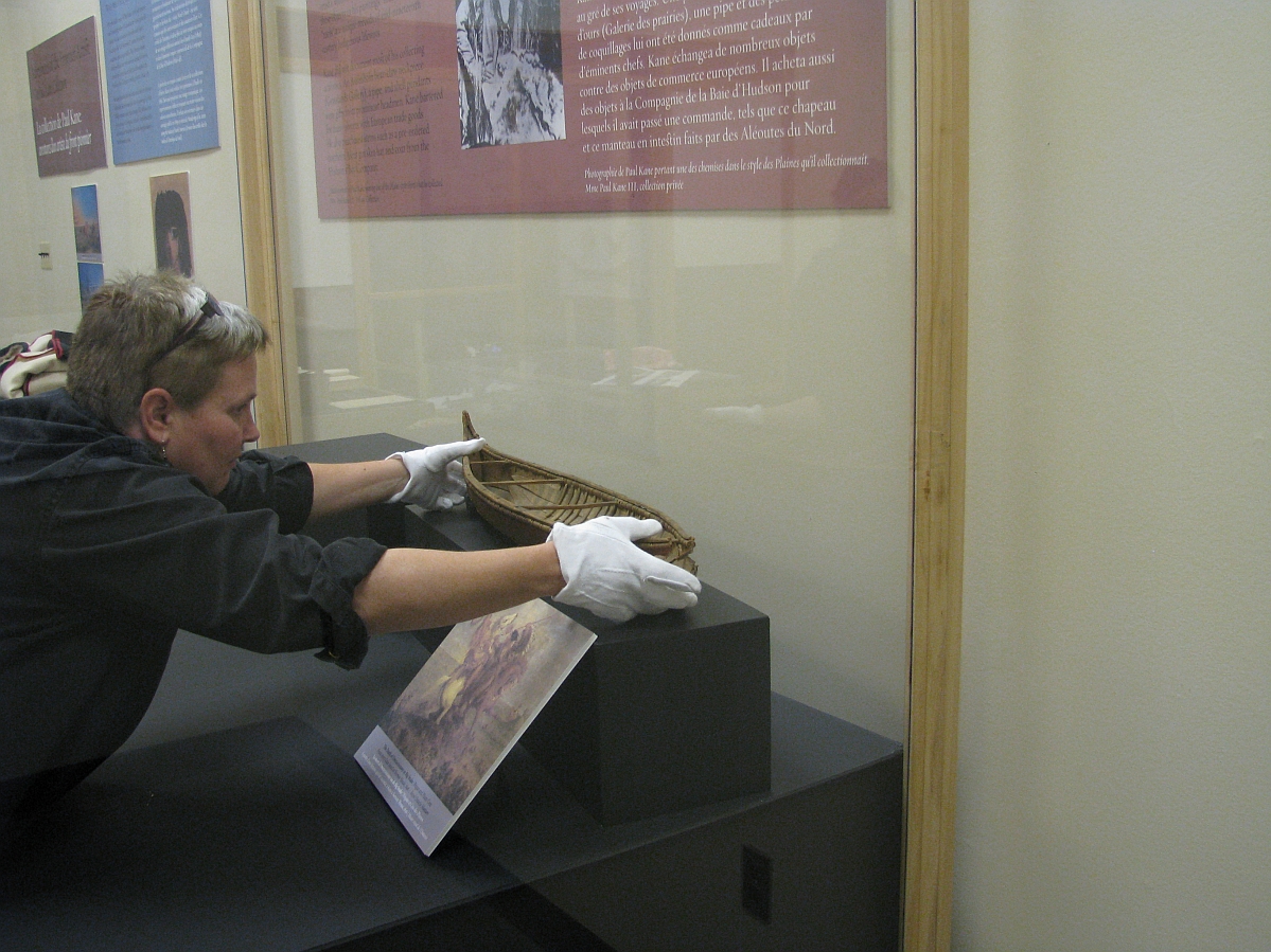 A museum staff person reaching into a display case, placing a model canoe in place.