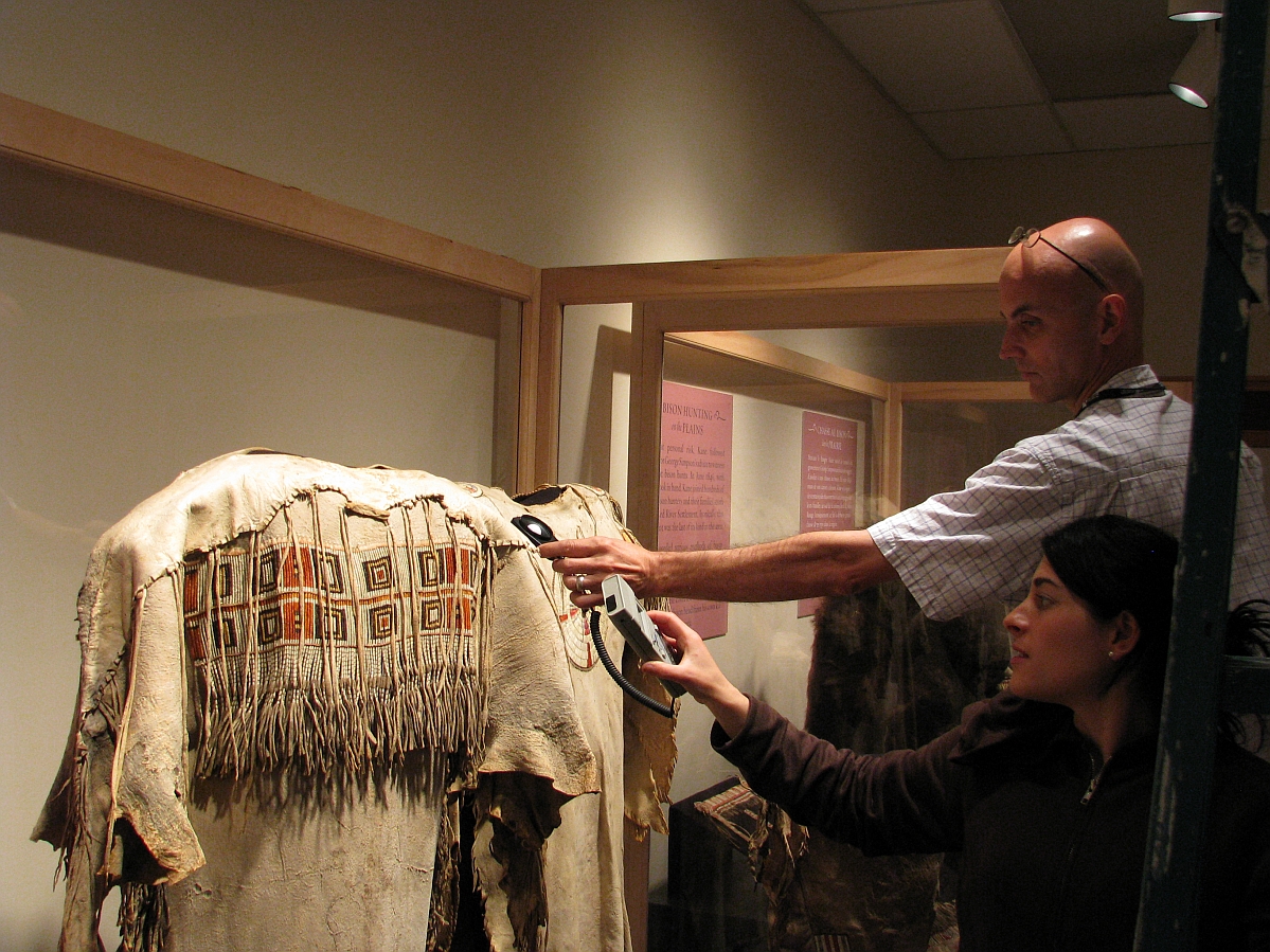 Two museum staff persons holding either end of a light reader over an object in a display case.