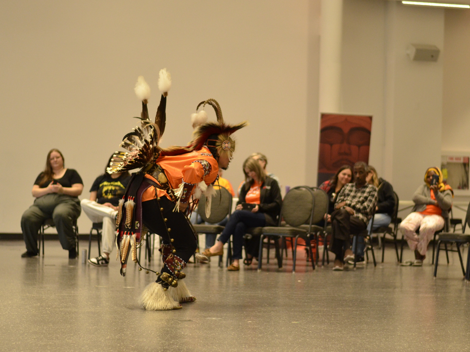 A dancer in traditional Indigenous regalia demonstrating a powwow dance for a seated crowd.