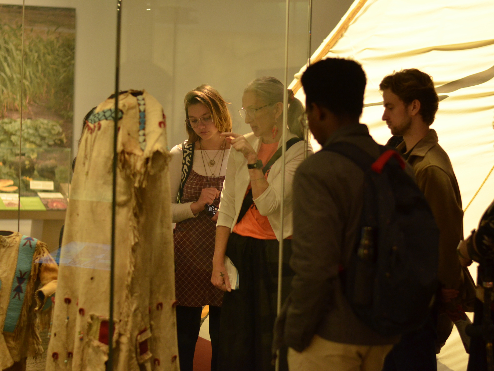 An older adult wearing an orange shirt speaking with a small group of younger adults in front of a display case containing an example of traditional Indigenous clothing.