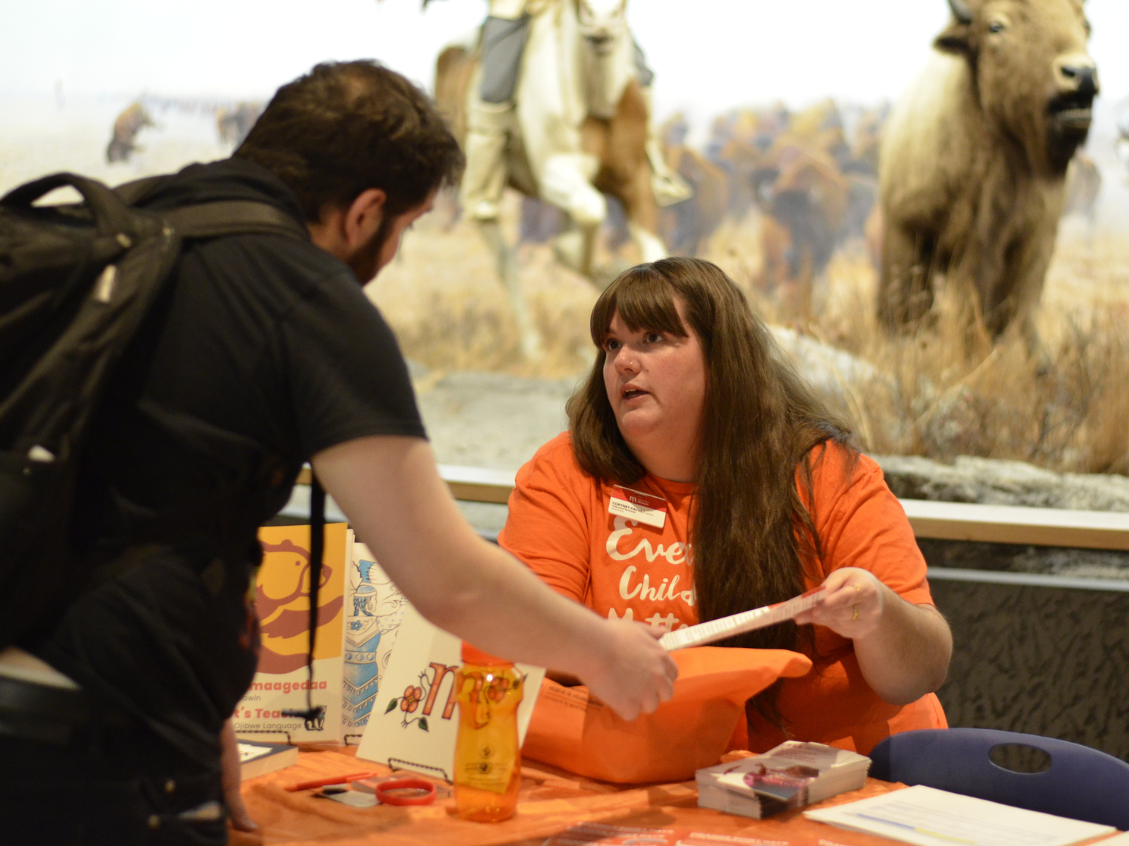 A Museum staff person wearing an orange shirt and seated at a table with an orange tablecloth shows something on a piece of paper to a Museum visitor.