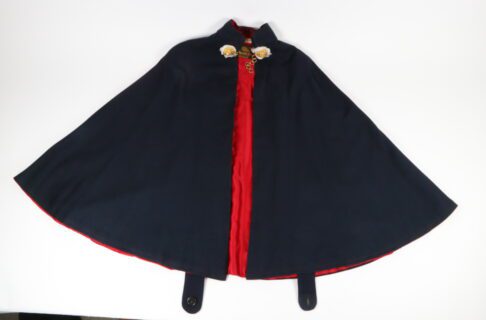 A black or dark navy cape with a red silk lining laid out on a white surface. At the collar is a gold chain and fasteners.