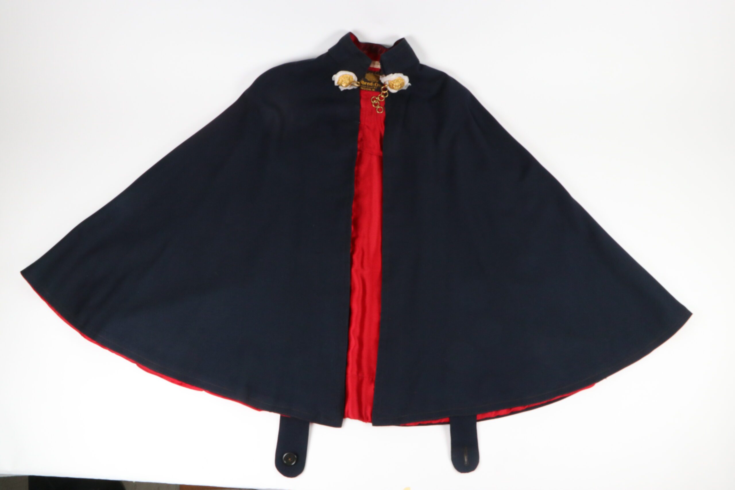 A black or dark navy cape with a red silk lining laid out on a white surface. At the collar is a gold chain and fasteners.