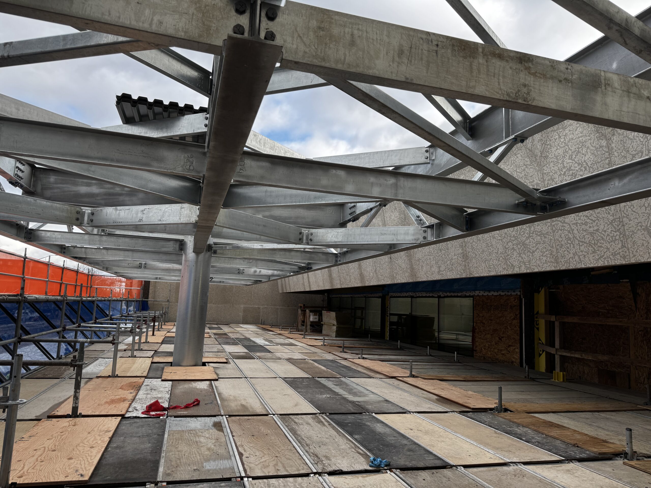 Looking out over the scaffolding level beneath the metal framing of the new awning over the Rupert Avenue entrance. The sky can be seen through criss-crossing metal beams over a boarded surface.