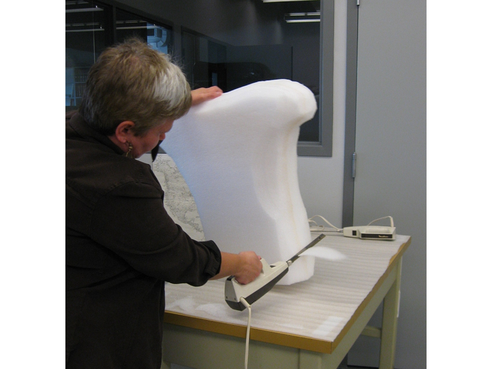 An individual using an electric knife hand tool shaping a large piece of foam on a table top.