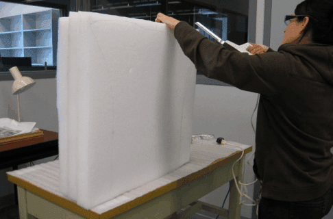 An individual using a hand tool to carve off a section of a large square of foam on a table.