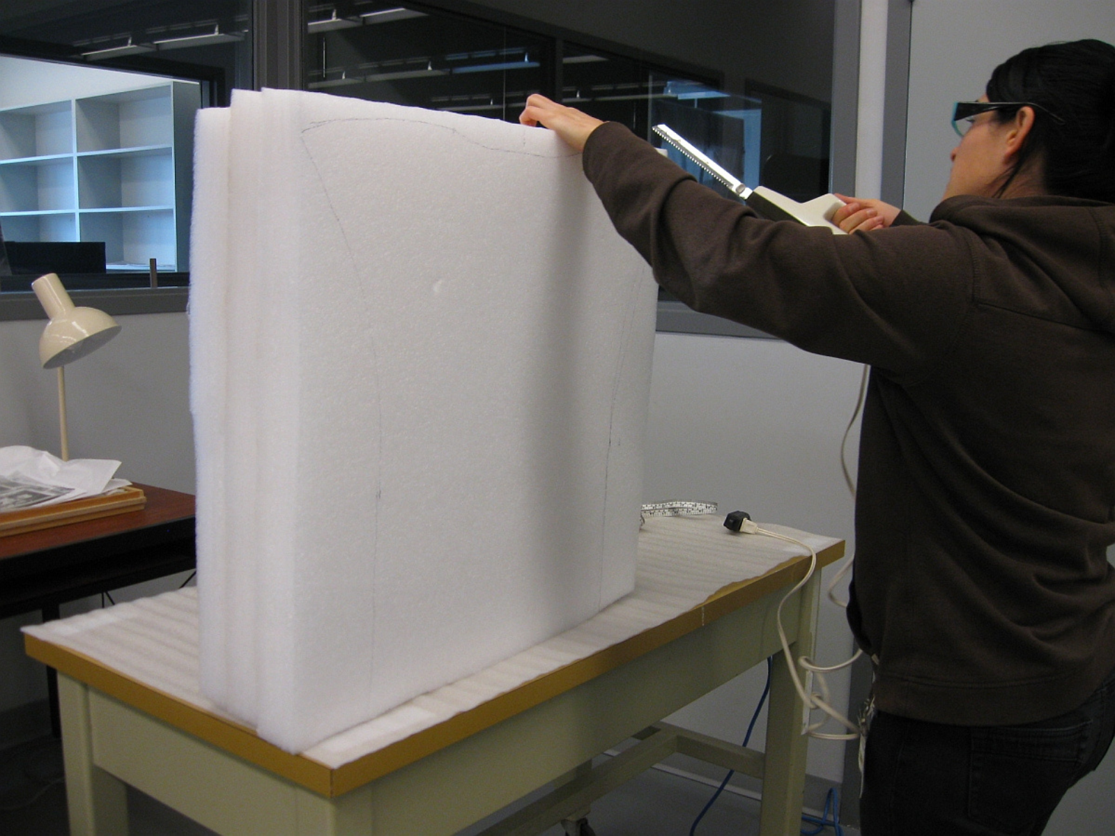 An individual using a hand tool to carve off a section of a large square of foam on a table.