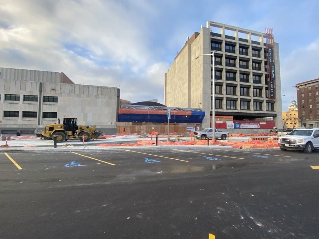 Photograph looking at the Manitoba Museum Rupert entrance from the parking lot. The Rupert entrance and Rupert Ave. are closed and under construction, with a large yellow digger driving down the closed road. Four handicapped parking spots are designated by painted symbols in a newly paved parking lot.