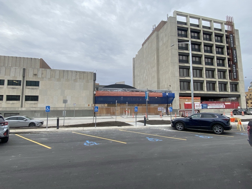 Photograph looking at the Manitoba Museum Rupert entrance from the parking lot. The Museum's Rupert entrance is closed and under construction. Four handicapped parking spots are designated by painted symbols in a newly paved parking lot.