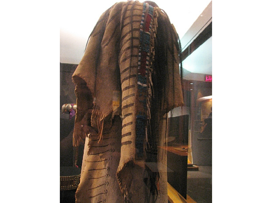 A hide shirt on a mannequin with elaborate beadwork going down the sleeve in a display case.