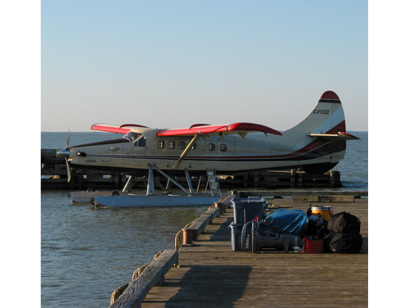 A float plane on the water near a dock, on which there's a pile gear and a boat motor.