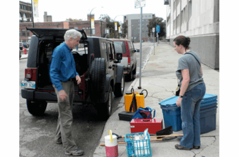 Two individuals with a variety of packs and bins of gear on the sidewalk next to a Jeep with an open back hatch.