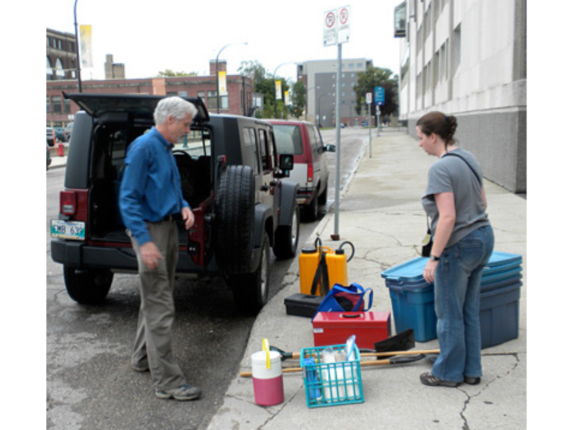 Two individuals with a variety of packs and bins of gear on the sidewalk next to a Jeep with an open back hatch.