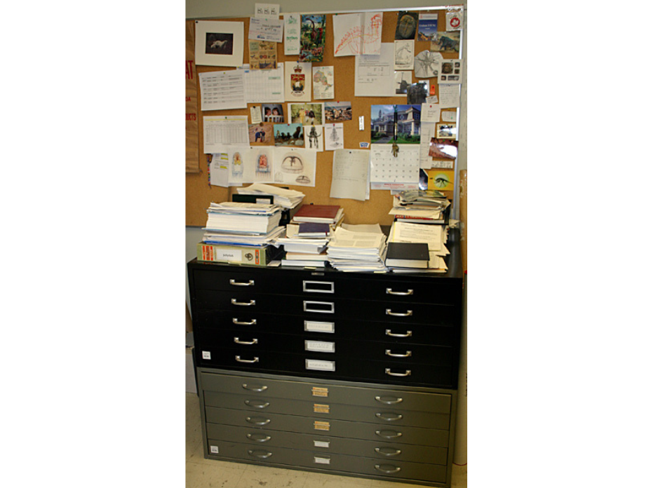 Two stacked cabinet with thin drawers against a wall below bulletin board filled with pinned papers and images.
