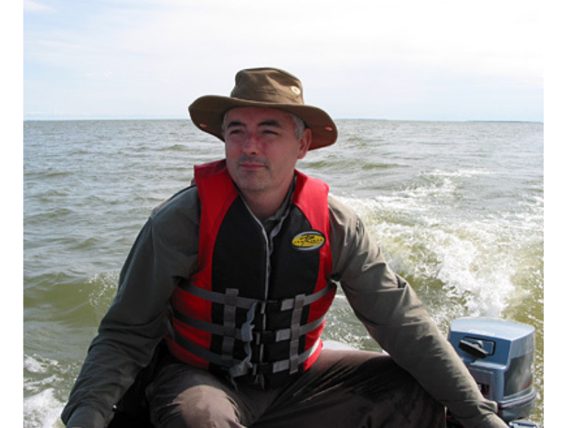 An individual wearing a brimmed hat and a red and black lifejacket steering the motor of a boat on a lake.
