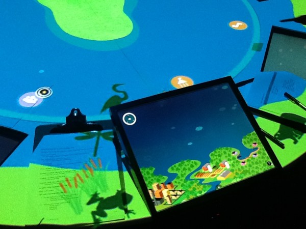 A brightly illuminated section of a rounded table with a digital touchscreen embedded in the surface. On the table itself and the touchscreen, illustrations of a lake and small creatures like frogs, herons, and fish.