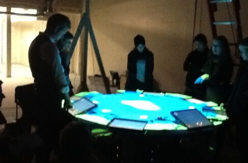 Six people stand around a brightly illuminated round table in a dim room.
