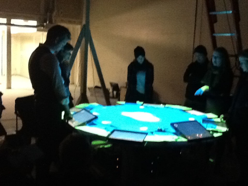Six people stand around a brightly illuminated round table in a dim room.