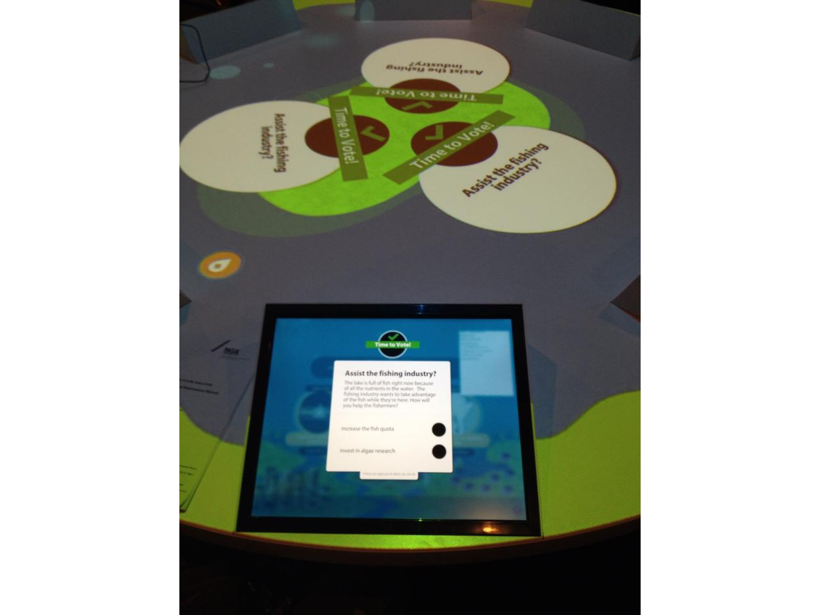 Looking at a touchscreen embedded in a round table., In the centre of the table projections ask the vote question "Assist the fishing industry?". The screen shows the same question with selectable options to "Increase fish quota" or "Invest in algae research.