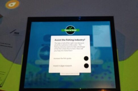 Looking at a touchscreen embedded in a round table. A pop-up on the screen asks the vote question "Assist the fishing industry?" followed by selectable options to "Increase fish quota" or "Invest in algae research.