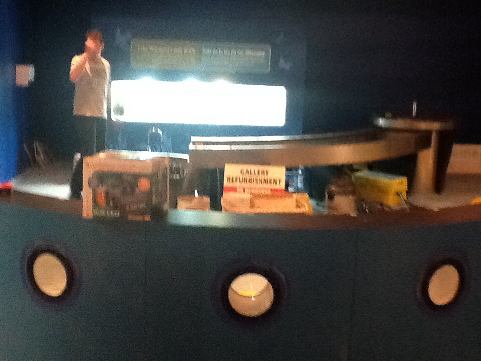 A grainy photo looking over a low half wall with three porthole windows towards a sloping watershed table, and a brightly illuminated rectangle section of wall where a aquarium tank will be installed.