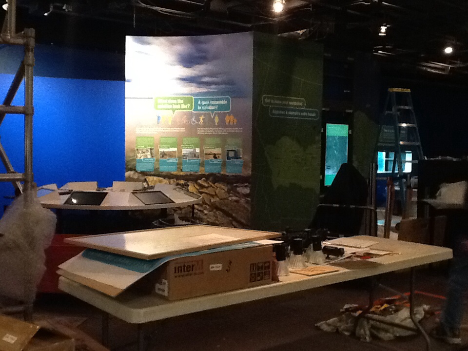 An exhiit under construction in the Manitoba Museum Science Gallery. A table holds boxes, papers, and tools in the foreground.